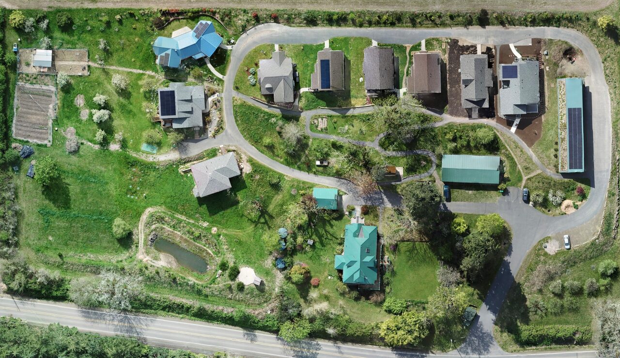 Aerial view of the property showing nine houses along a single loop road, the common buildings, garden beds, orchard rows, the pond and open lawns.