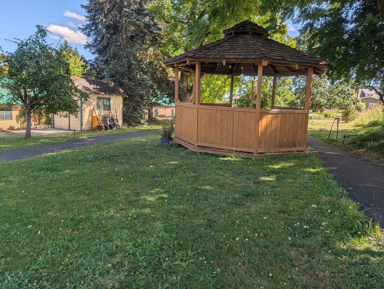 A wooden gazebo on a mown lawn, with a paved path running past it toward the houses.